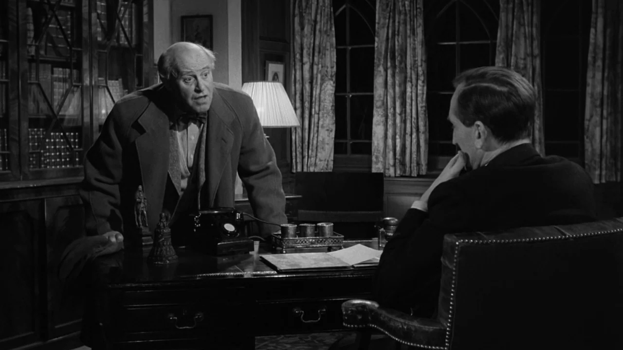Village of the Damned (1960) movie still: medium — A wood-paneled room features a desk cluttered with old documents and a rotary phone. One man…