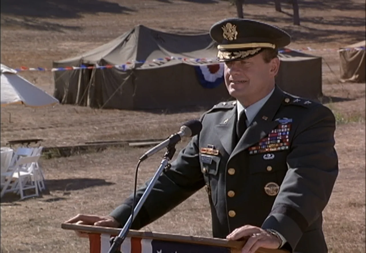 The Pentagon Wars (1998) movie still: medium — An outdoor military event takes place on a dusty dirt field with canvas tents. A high-ranking officer…