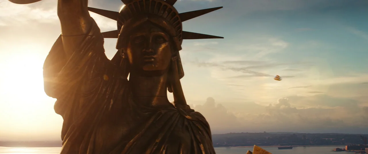 The Marvels (2023) movie still: medium — The Statue of Liberty is illuminated by the warm light of a golden sunset. A small golden craft flies…
