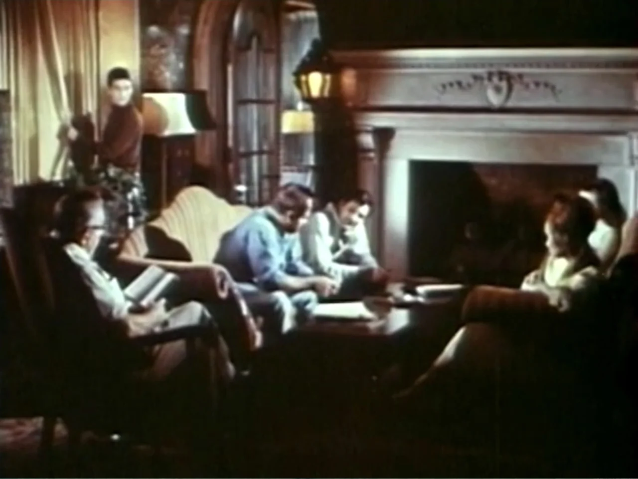 In the Year 2889 (1969) movie still: medium — A living room features a large stone fireplace and traditional upholstered furniture. Four people sit…
