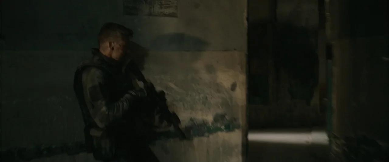 Hounds of War (2024) movie still: medium — A dark, dilapidated hallway with peeling paint and exposed concrete surfaces. A soldier in tactical gear…
