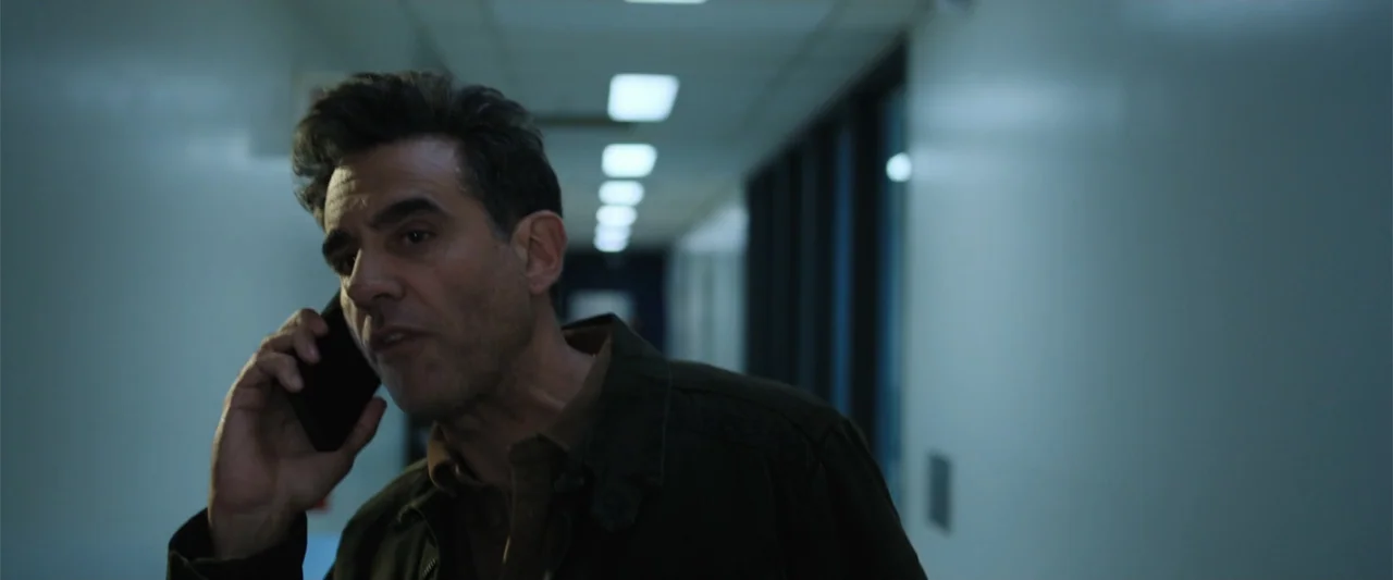Trap House (2025) movie still: medium — An interior hallway features white walls and a ceiling with rows of recessed lights. A middle-aged man…
