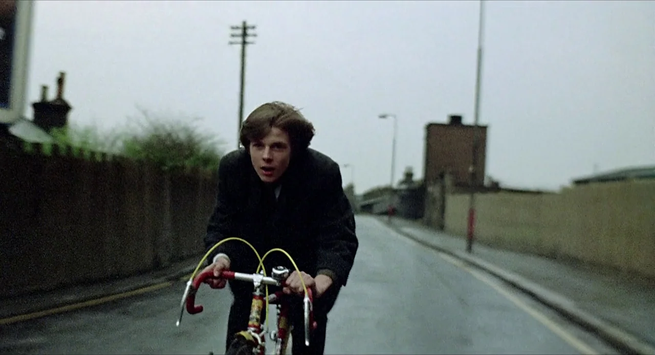 Deep End (1970) movie still: medium — A quiet paved suburban road is flanked by stone walls and utility poles under overcast skies. A young man…