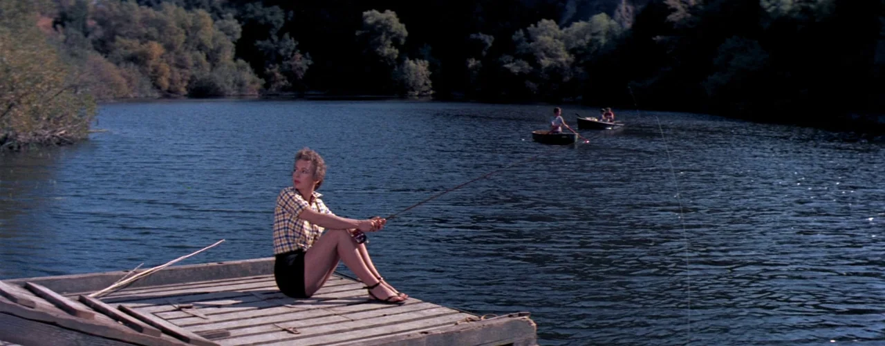 The Seven Year Itch (1955) movie still: wide — A wooden raft floats on calm water near a wooded shoreline under daylight. A woman in a plaid shirt…