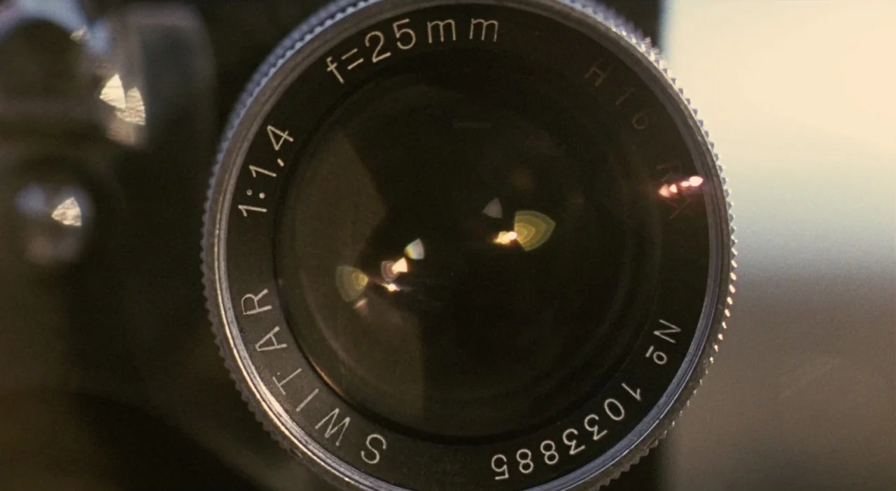 Patagonia (2010) movie still: closeup — A close-up view of a vintage Switar camera lens with silver metal housing. The black lens glass shows bright…