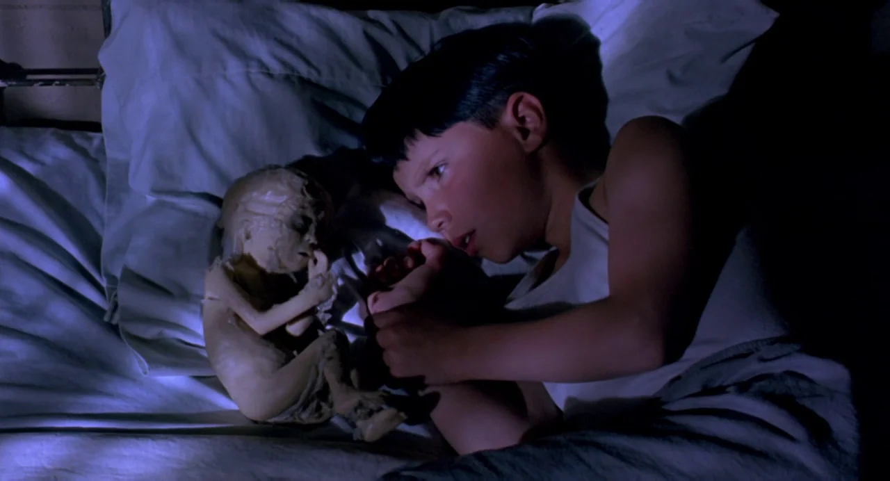 The Reflecting Skin (1990) movie still: medium — A dark, dimly lit indoor setting features a child resting on a soft bed. The child gazes at a…
