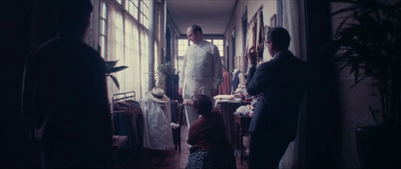 Neruda (2016) movie still: fullbody — A long narrow room with large windows features a clothing alteration in progress. An older man stands for…