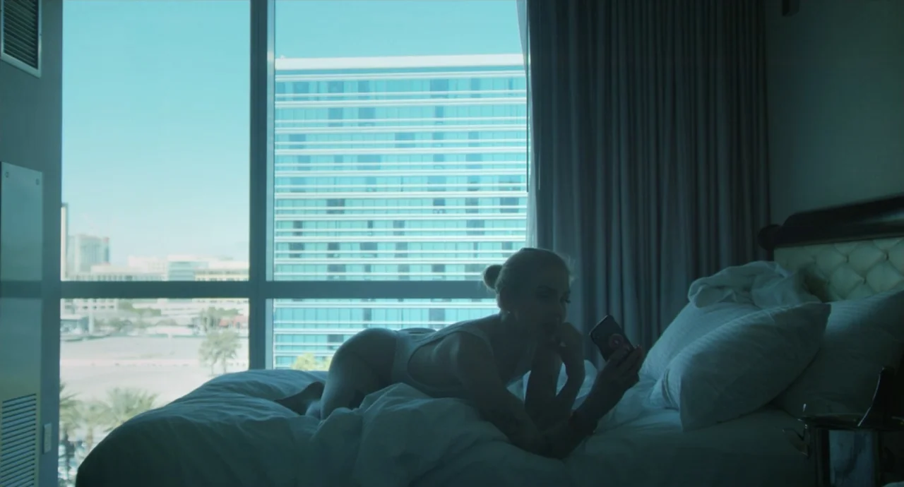 Pleasure (2021) movie still: medium — A high-rise hotel room features a large window overlooking a modern urban complex. A woman in a white bodysuit…