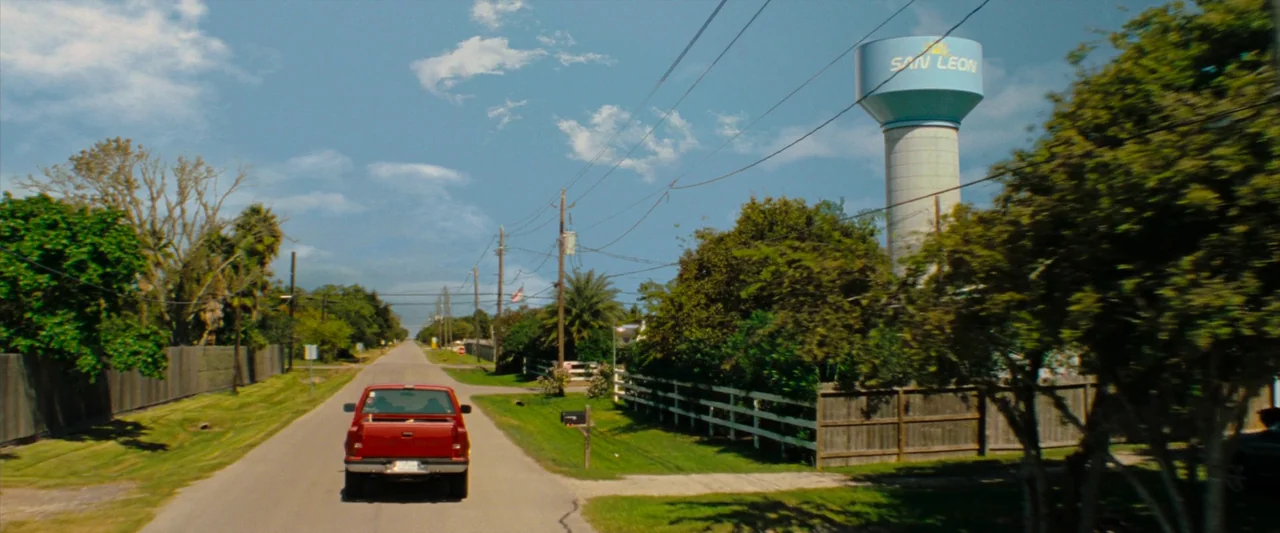 Red Rocket (2021) movie still: wide — A suburban road features a water tower marked San Leon against a clear sky. A red pickup truck travels away…