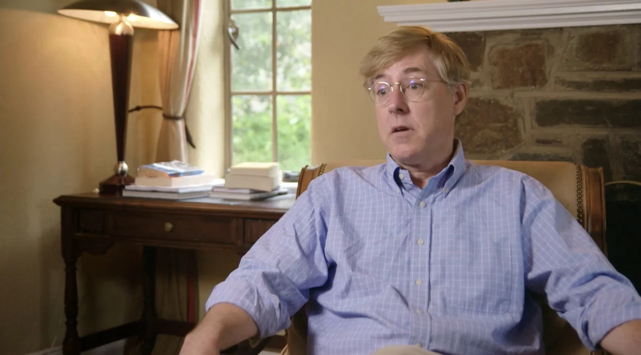 The Divided Soul of America (2019) movie still: medium — A man sits in an armchair inside a room with stone fireplace and desk. He wears a blue…