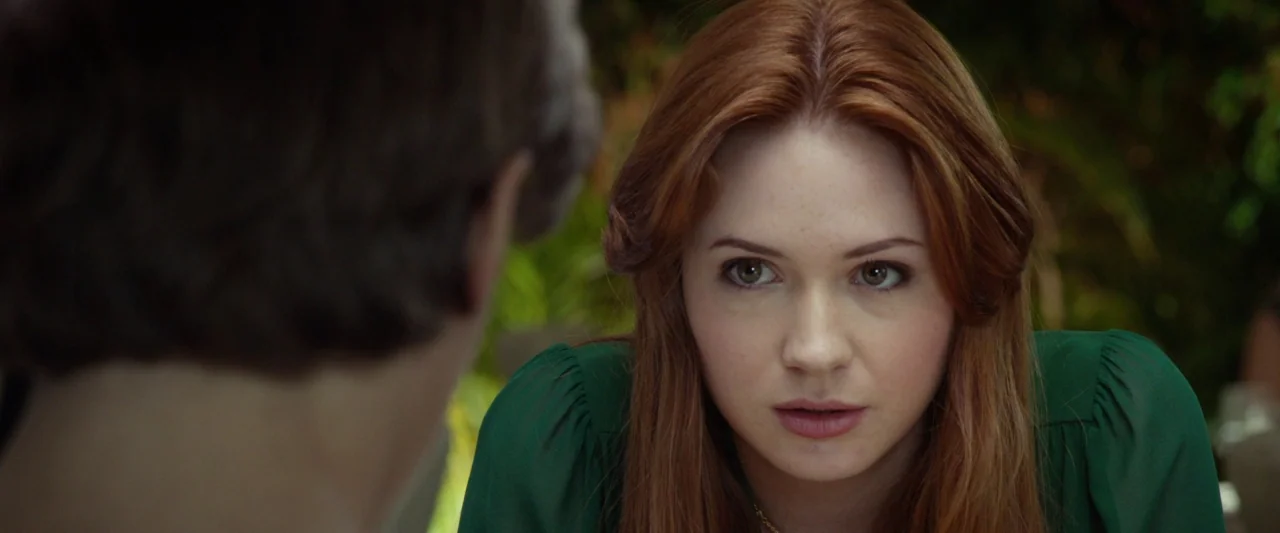 Oculus (2013) movie still: closeup — A woman sits outdoors in a garden area filled with out-of-focus green plants. She faces an unseen person whose…