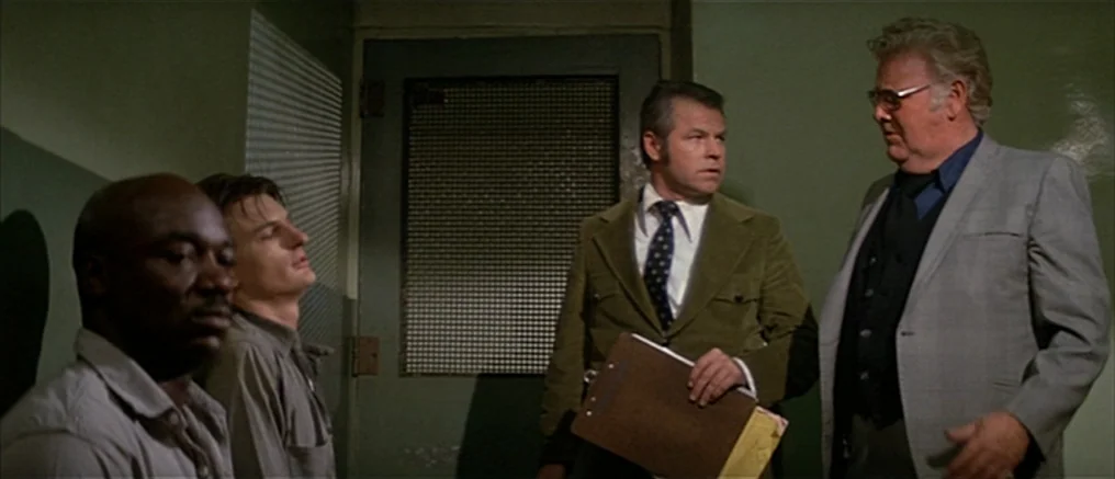 Assault on Precinct 13 (1976) movie still: medium — A small room with light green walls features a heavy door with wire mesh. Two men in suits stand…