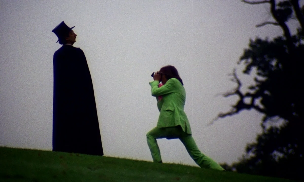 Wonderwall (1968) movie still: fullbody — Two men stand on a grassy slope under a bright overcast sky. One man in a lime green suit photographs a…