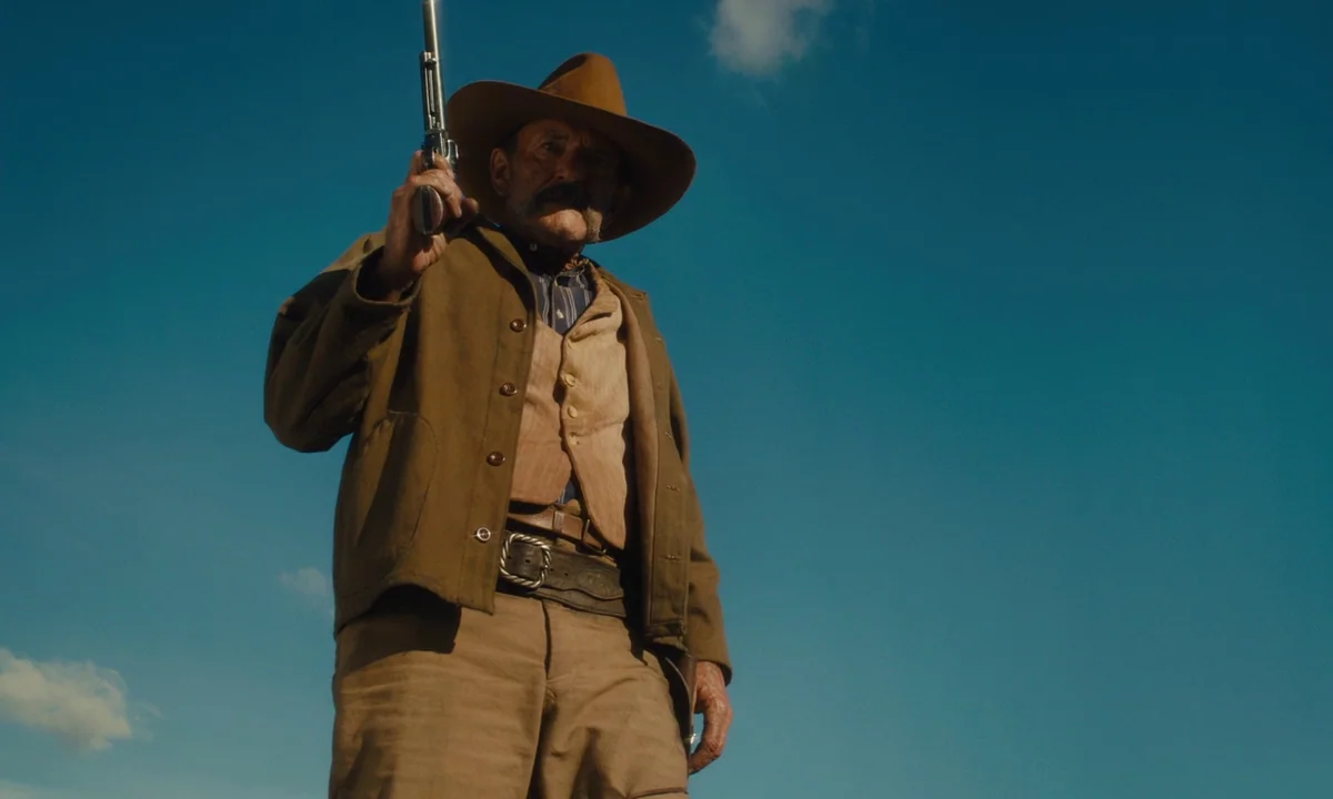 Slow West (2015) movie still: medium — An open landscape with a clear blue sky dominates the frame. An elderly cowboy stands holding a revolver…