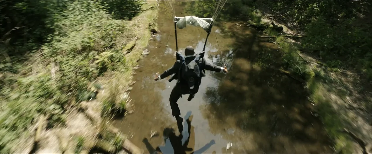 Heads of State (2025) movie still: wide — A man descends by parachute into a narrow creek bed surrounded by dense foliage. He is suspended over the…