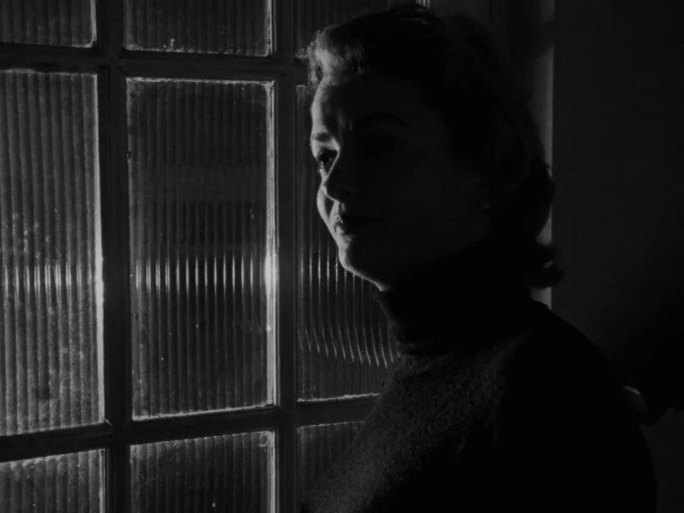 Following (1998) movie still: medium — A dimly lit interior space features a woman standing beside a textured glass window. She gazes out into the…