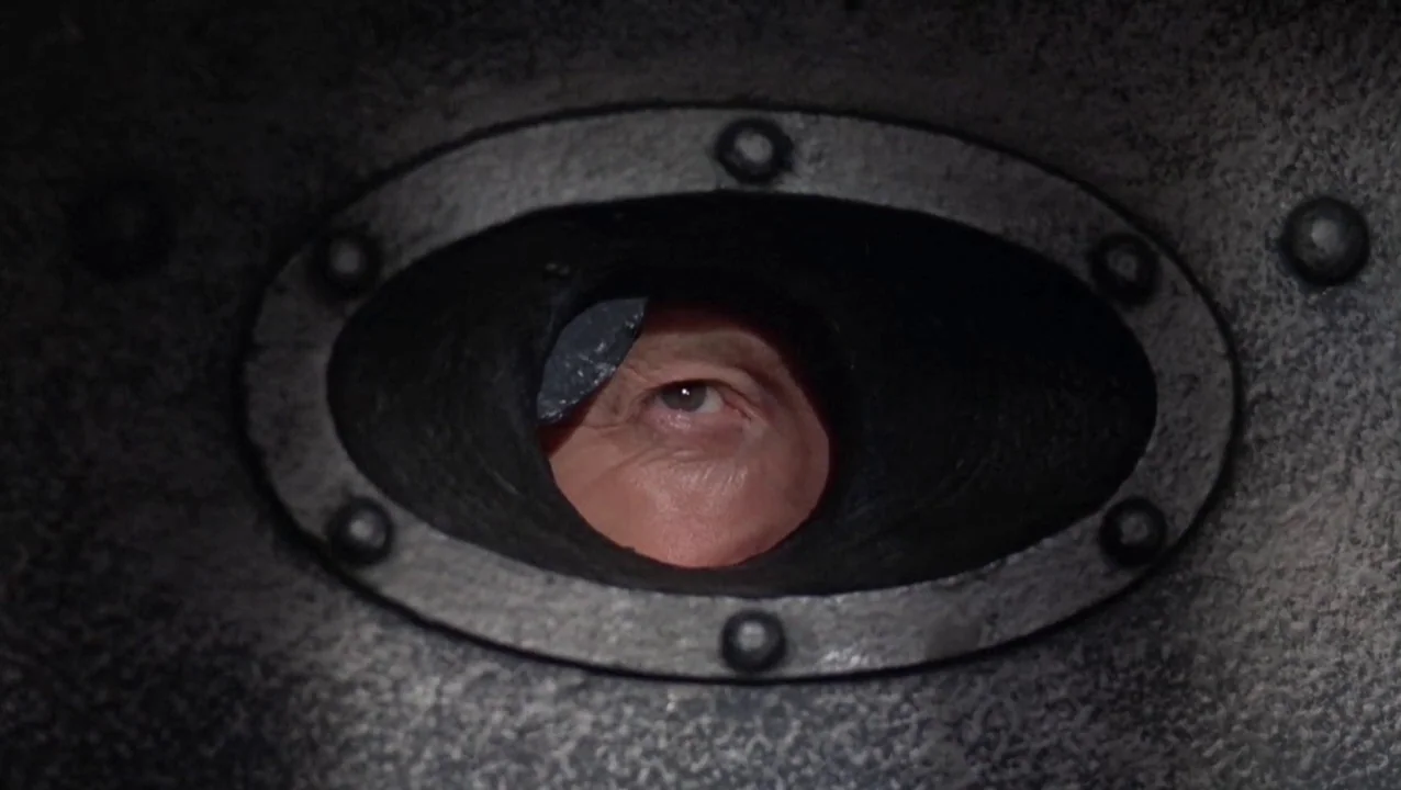 O Lucky Man! (1973) movie still: closeup — A heavy metal surface features a small, oval aperture secured with iron rivets. A human eye gazes…