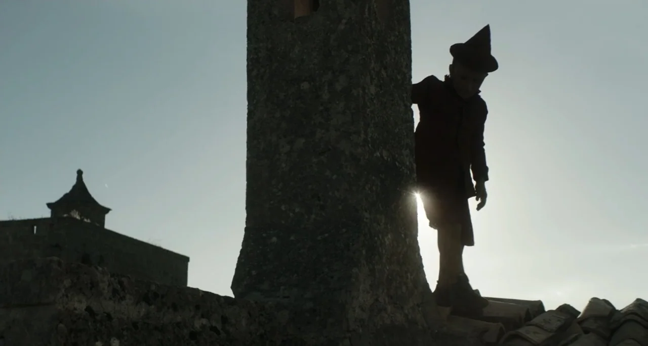Pinocchio (2022) movie still: fullbody — A stone rooftop features a large chimney and a tiled surface in bright daylight. A young boy wearing a…