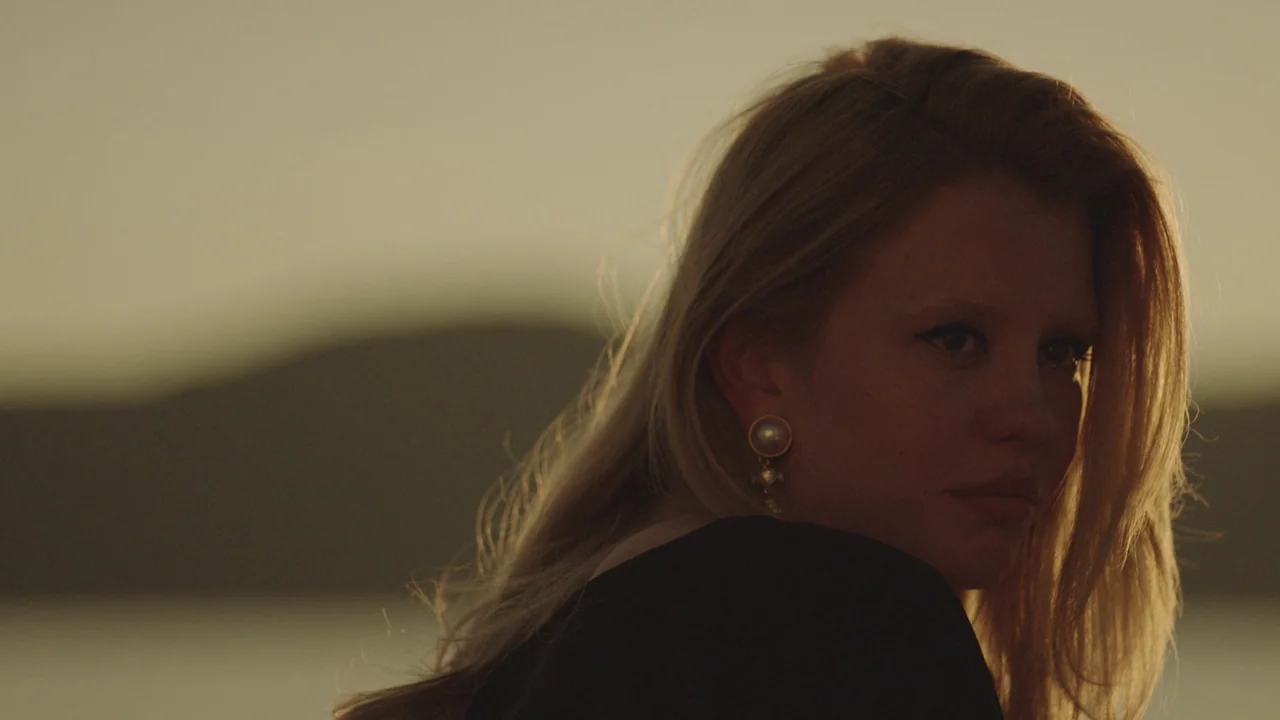 Infinity Pool (2023) movie still: closeup — A woman stands near a coastal location during the low golden light of dusk. The subject turns her head…