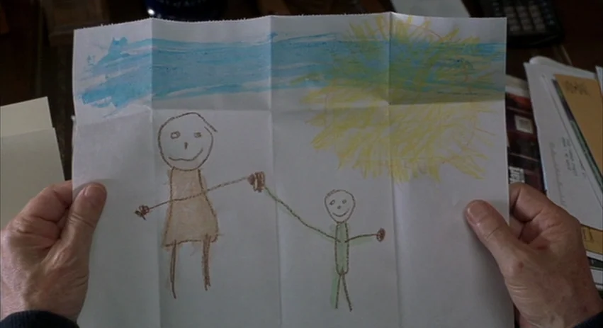 About Schmidt movie still: closeup — A pair of wrinkled hands hold a folded sheet of white paper. The paper features a child's crayon drawing of two…