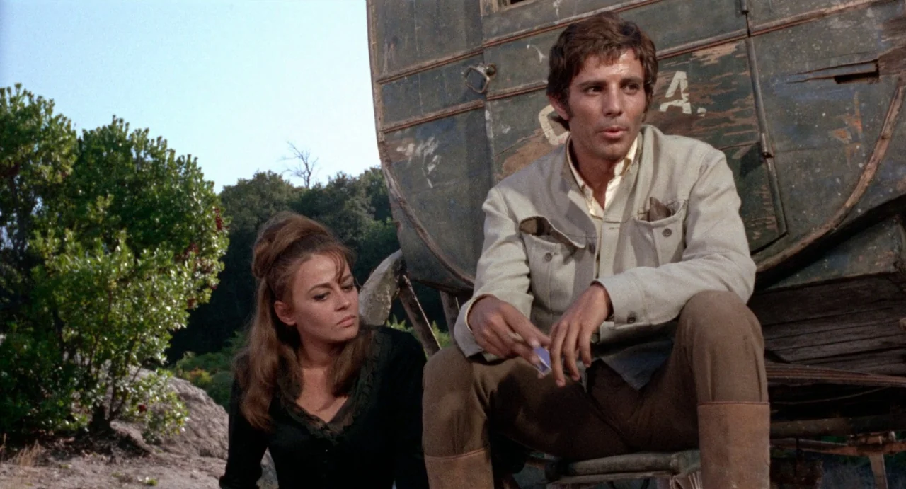 The Hellbenders (1967) movie still: medium — A man and woman sit next to an old wooden cart in a rural setting. The man holds a small object while…