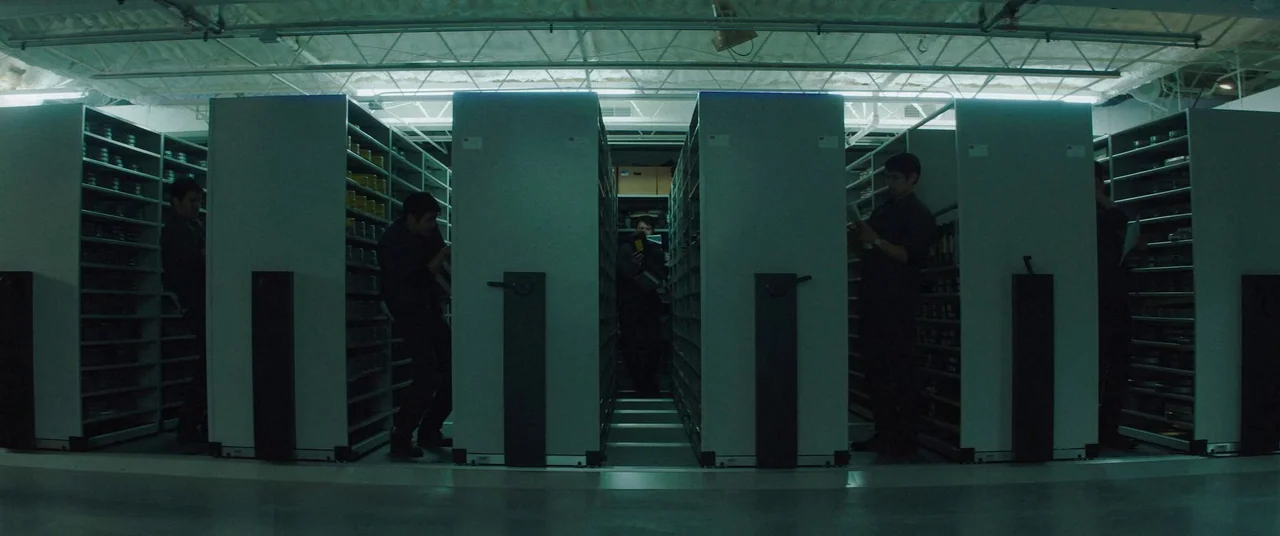 Broadcast Signal Intrusion (2021) movie still: wide — A rows of high-density mobile steel storage shelves stand in an industrial basement space.…