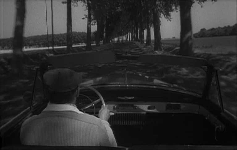 Le Signe du Lion (1962) movie still: medium — A convertible travels down a long road flanked by lines of tall, dense trees. The driver holds the…