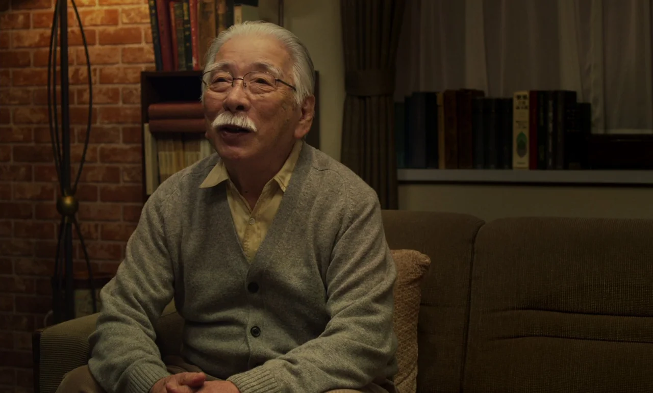 Like Someone In Love (2012) movie still: medium — An elderly man sits on a brown sofa in a home interior. Behind him are bookshelves and a brick…