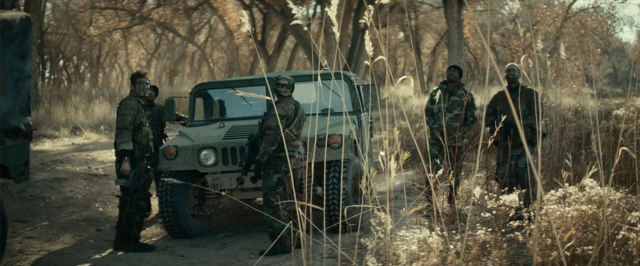 12 Strong (2018) movie still: wide — A group of soldiers stands on a dirt path in a wooded area. They are positioned around a green military humvee…
