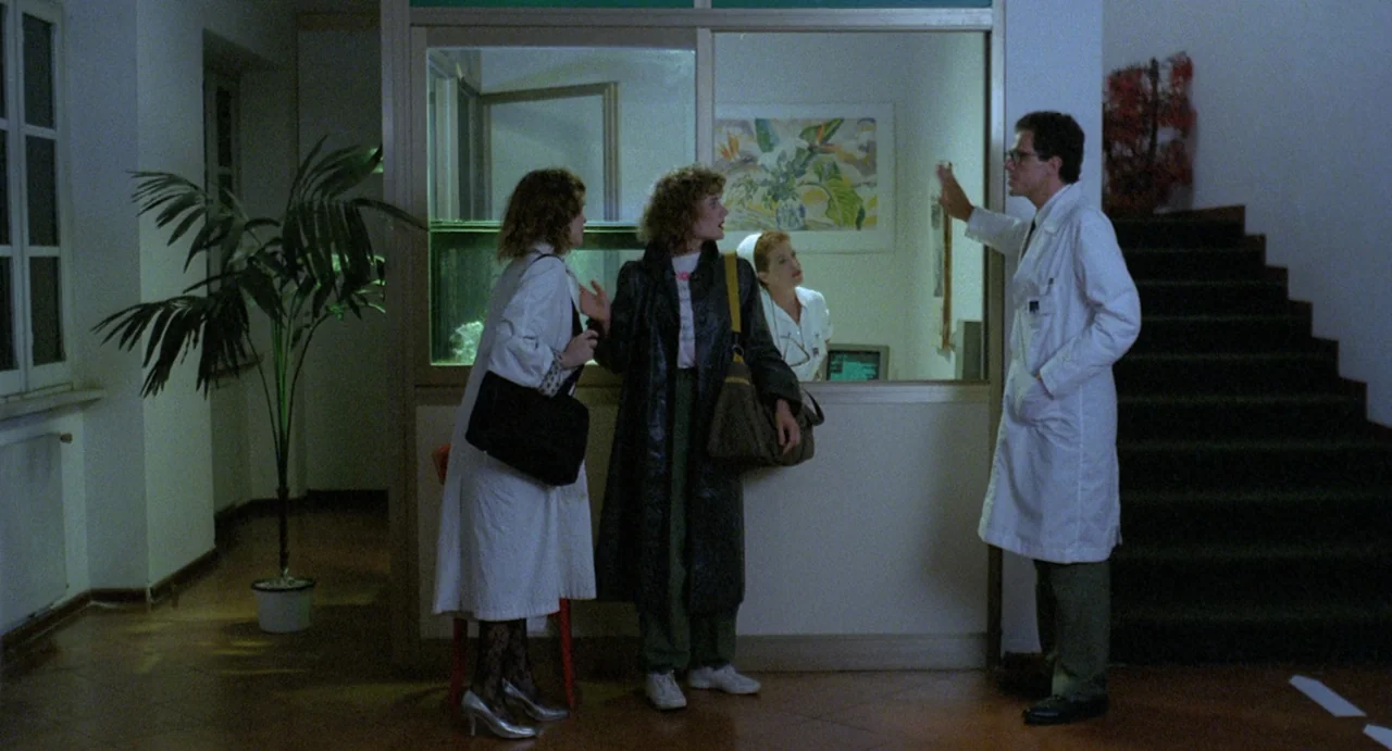 StageFright (1987) movie still: fullbody — A hospital lobby features a glass partition separating the public area from staff quarters. Three people…