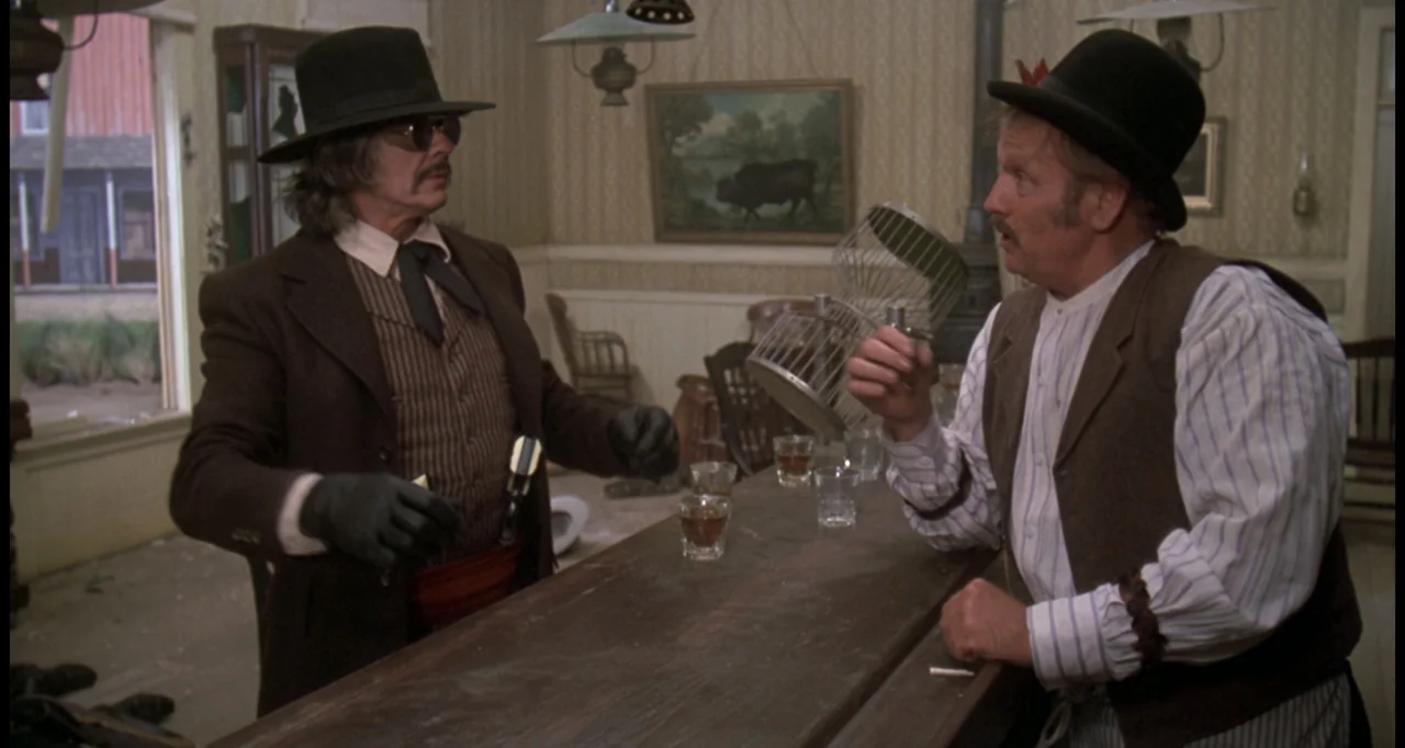 The White Buffalo (1977) movie still: medium — An 1800s western saloon has a long wooden bar and framed landscape paintings. Two men stand at the…