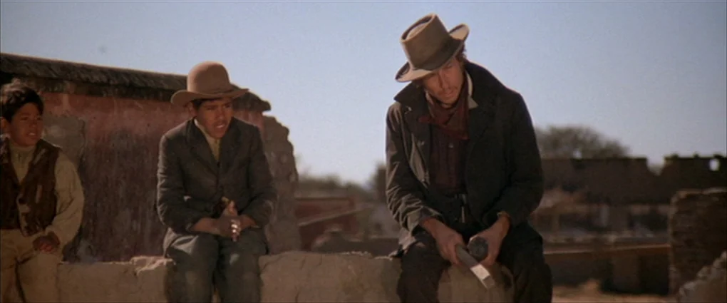 Pat Garrett & Billy The Kid (1973) movie still: medium — Three individuals sit atop a crumbling adobe wall in a dry, sparse landscape. The men are…