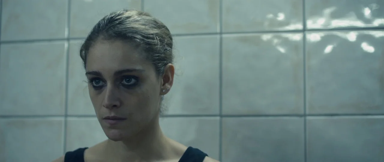 Alps (2011) movie still: closeup — A woman stands in front of light-colored grid-patterned ceramic wall tiles. She looks toward the left side of the…
