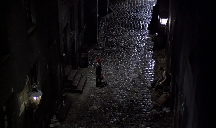 The Serpent’s Egg (1977) movie still: wide — A dark, narrow European-style cobblestone alleyway is illuminated by vintage streetlamps. Two men stand…