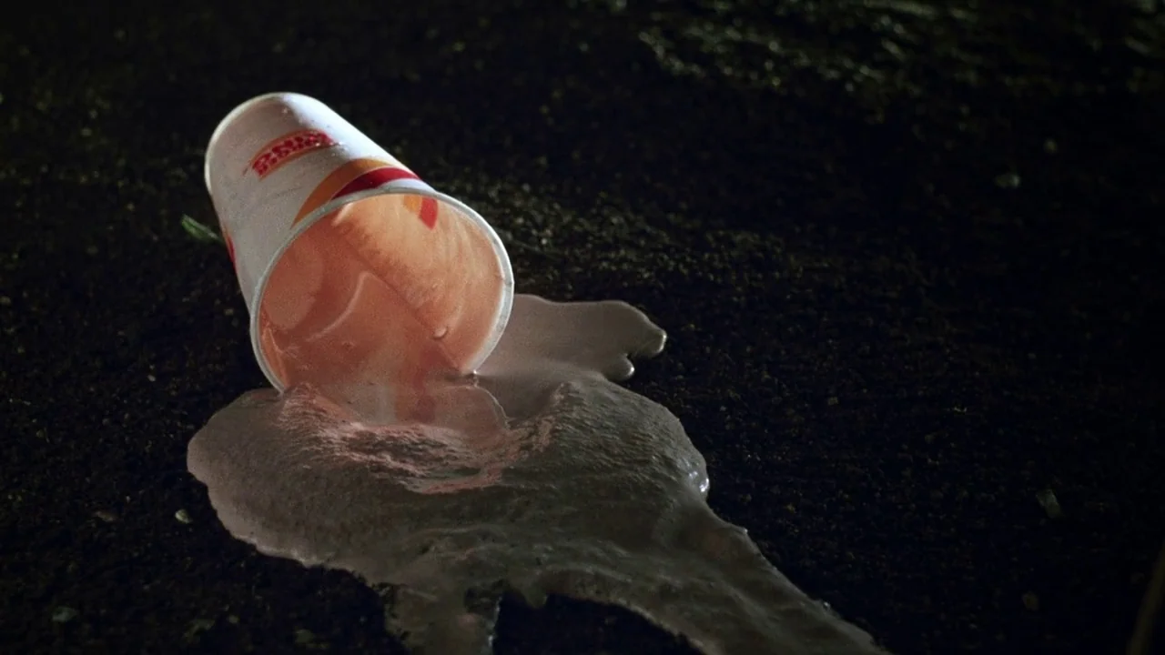 The Thin Blue Line (1988) movie still: closeup — A discarded fast food paper cup lies on a patch of dark, textured asphalt. A beige liquid contents…