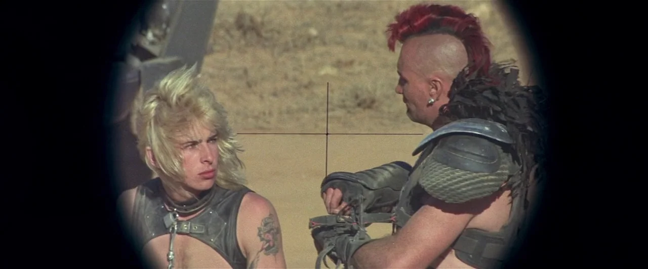 Mad Max 2 (1981) movie still: medium — A dry, sandy landscape serves as the backdrop for two post-apocalyptic warriors. The subjects are viewed…