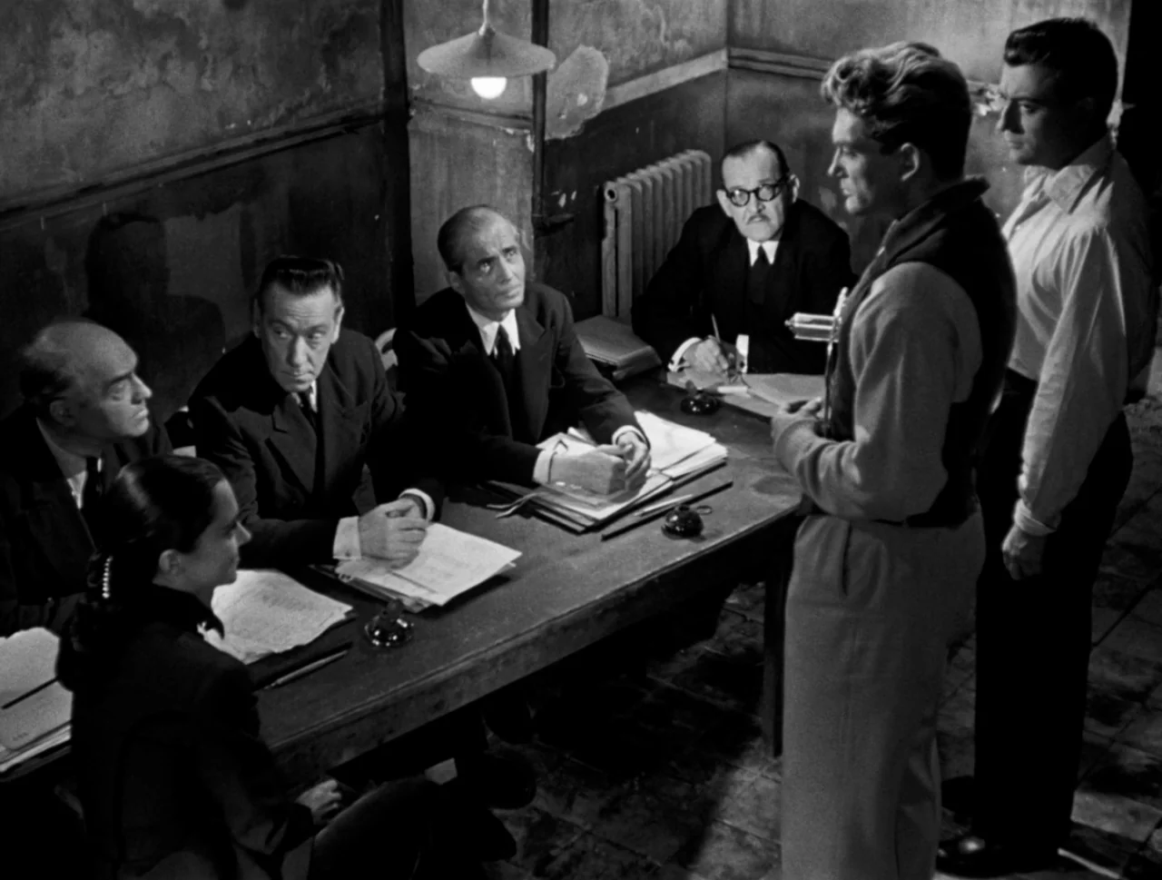 Orpheus (1950) movie still: medium — A dimly lit room contains six men and one woman gathered around a long table. The standing man addresses the…