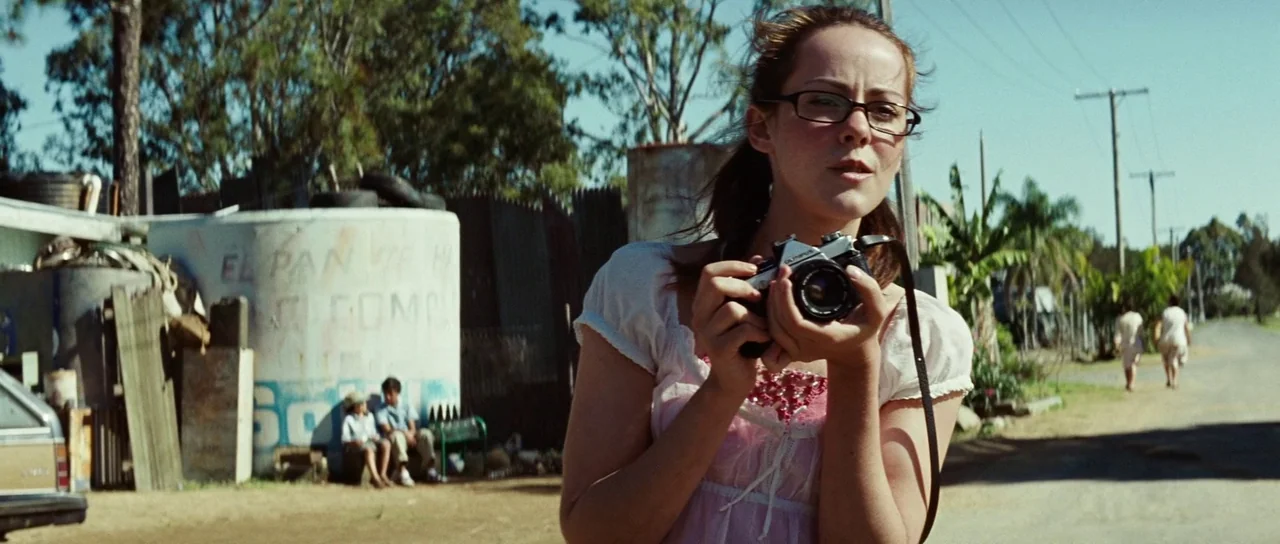 The Ruins (2008) movie still: medium — A rural, dusty roadside with a graffiti-covered water tank and scattered debris. A young woman in a lace top…