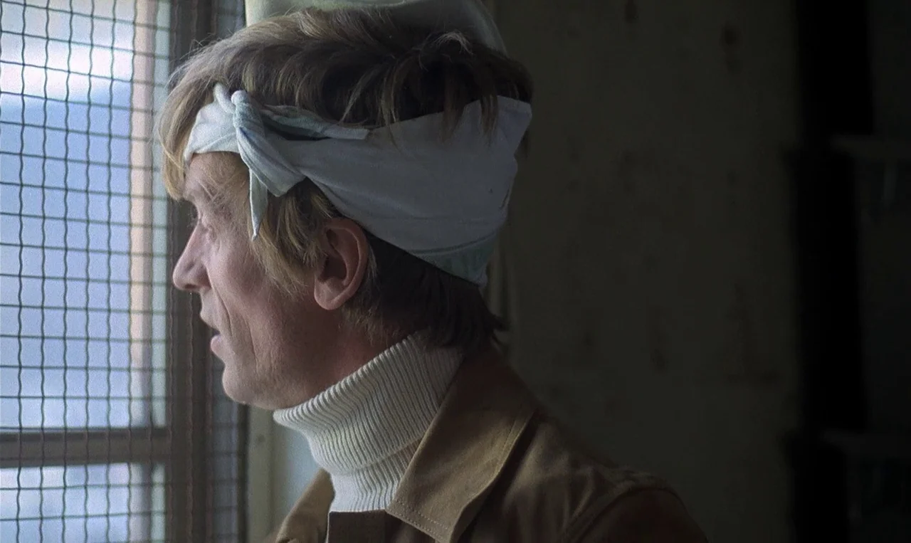 Man on the Roof (1976) movie still: closeup — A man stands in a dimly lit room beside a window covered with wire mesh. He wears a white head bandage…