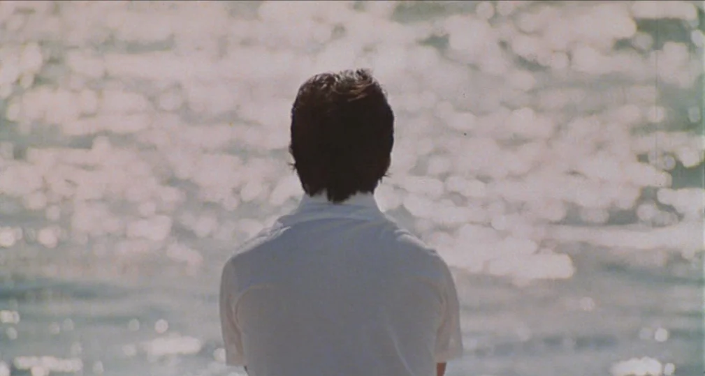 Midnight Cowboy (1969) movie still: medium — A person stands in front of a bright, shimmering expanse of water. The subject is seen from behind,…