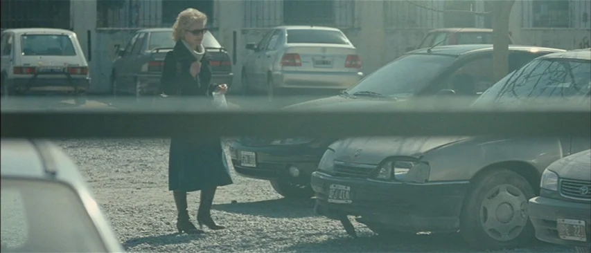 The Headless Woman (2008) movie still: medium — A gravel surface is crowded with numerous parked passenger vehicles on a bright day. A blonde woman…