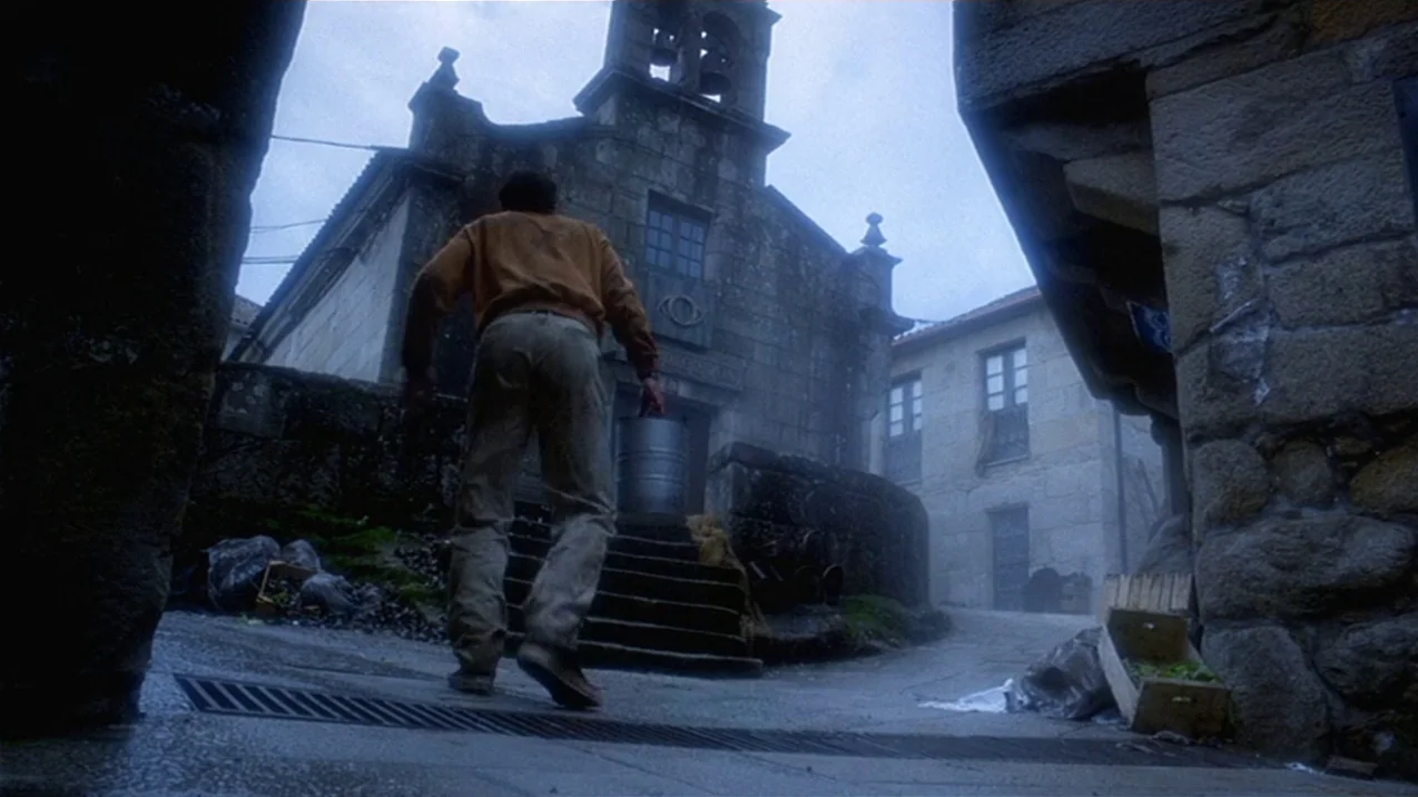 Dagon (2001) movie still: fullbody — A stone village plaza features an old church with an eye symbol above the door. A man carries a metal pail up…