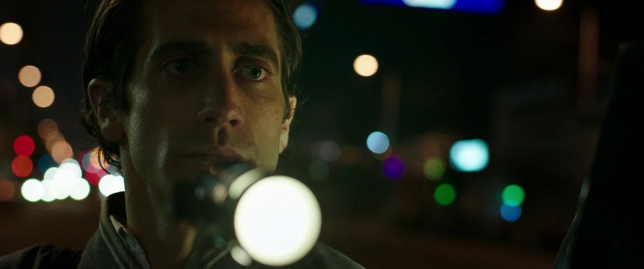 Nightcrawler (2014) movie still: closeup — A man stands on a city street at night surrounded by blurred traffic lights. He holds a bright circular…