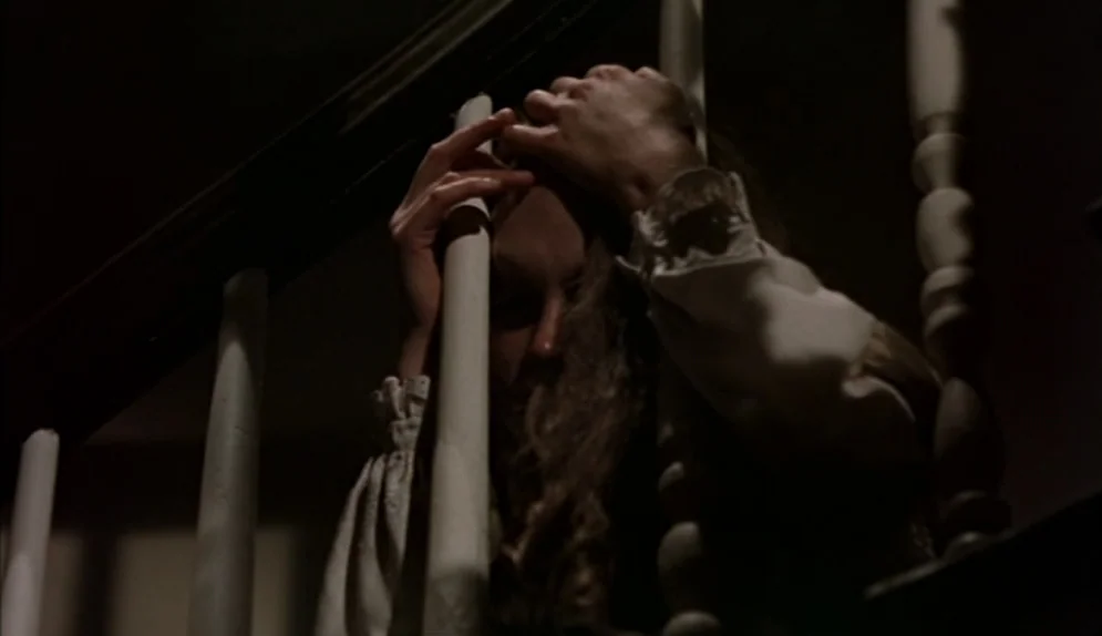The Beguiled (1971) movie still: closeup — A dark staircase interior featuring white wooden railings and shadows. A woman sits behind the balusters…