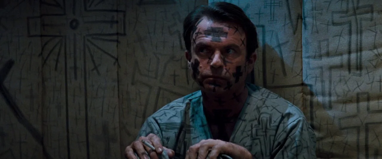 In The Mouth of Madness (1994) movie still: medium — A man sits inside a small space with walls covered in hand-drawn black crosses. The subject…