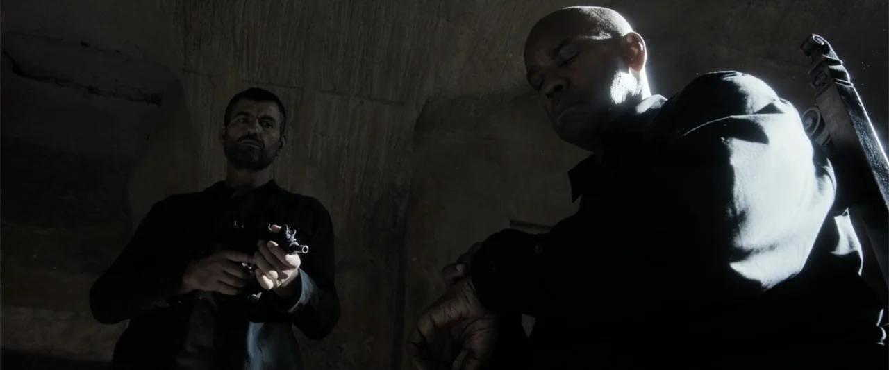 The Equalizer 3 (2023) movie still: medium — A stone basement room with dark shadows and curved architecture. Two men are present while one holds a…