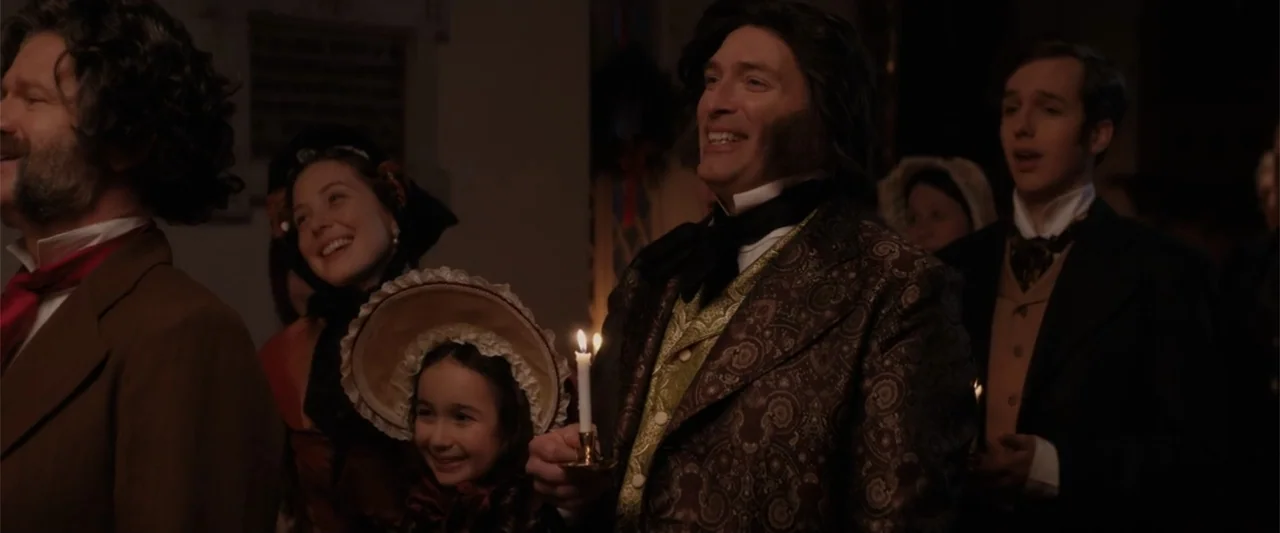 I Heard the Bells (2022) movie still: medium — A 19th-century church interior is dimly lit by the warm glow of handheld candles. A group of men,…