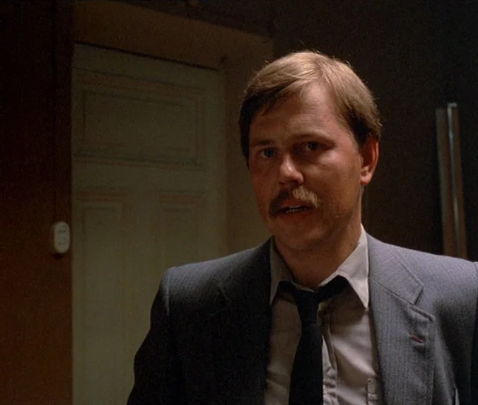 Crime & Punishment (1983) movie still: medium — An office interior features a wooden paneled wall and a white closed door. A man in a suit looks…