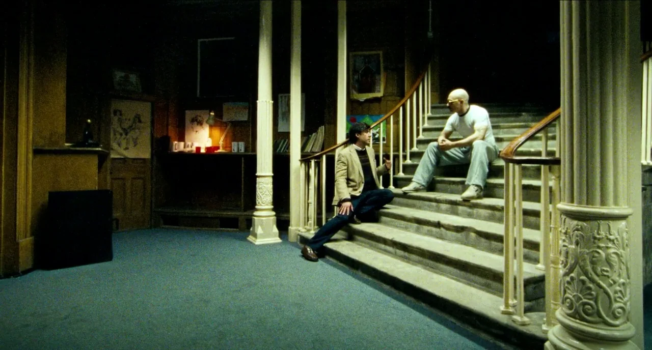 Bronson (2008) movie still: wide — A dimly lit grand staircase with ornate white pillars and a wooden handrail. Two men occupy the lower steps while…