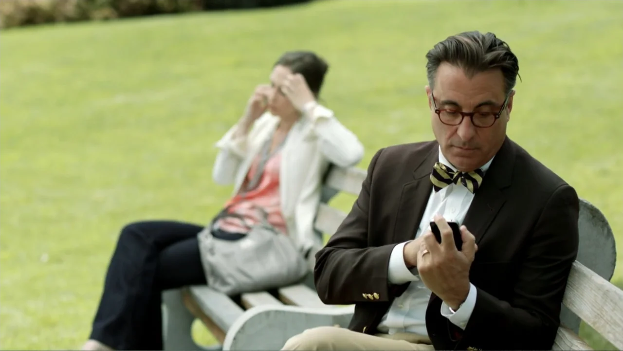 At Middleton (2013) movie still: medium — An outdoor park setting with a green lawn and white wooden benches. A man in a blazer sits in the…
